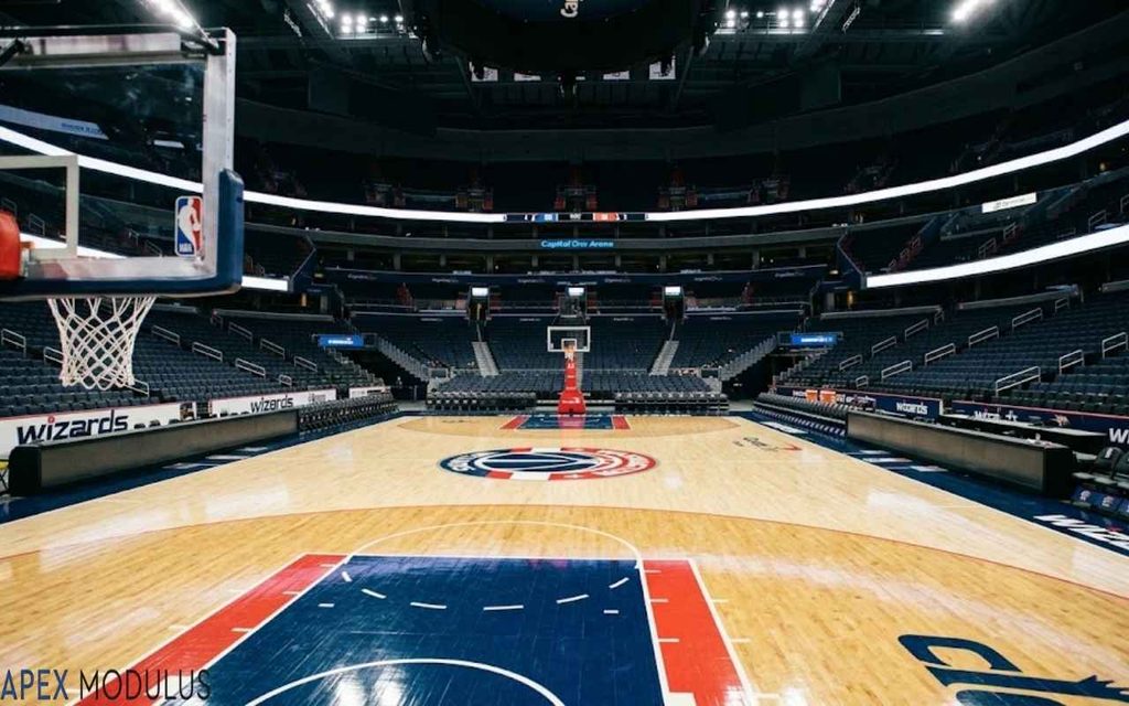 An empty 100 feet NBA basketball arena viewed from behind a hoop, looking down the polished hardwood court toward the opposing basket under bright overhead arena lights.