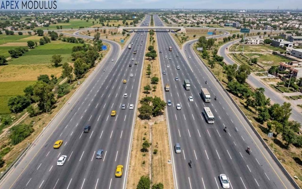 An aerial view looking down at a multi-lane highway with moderate traffic, flanked by green agricultural fields on one side and a town on the other.