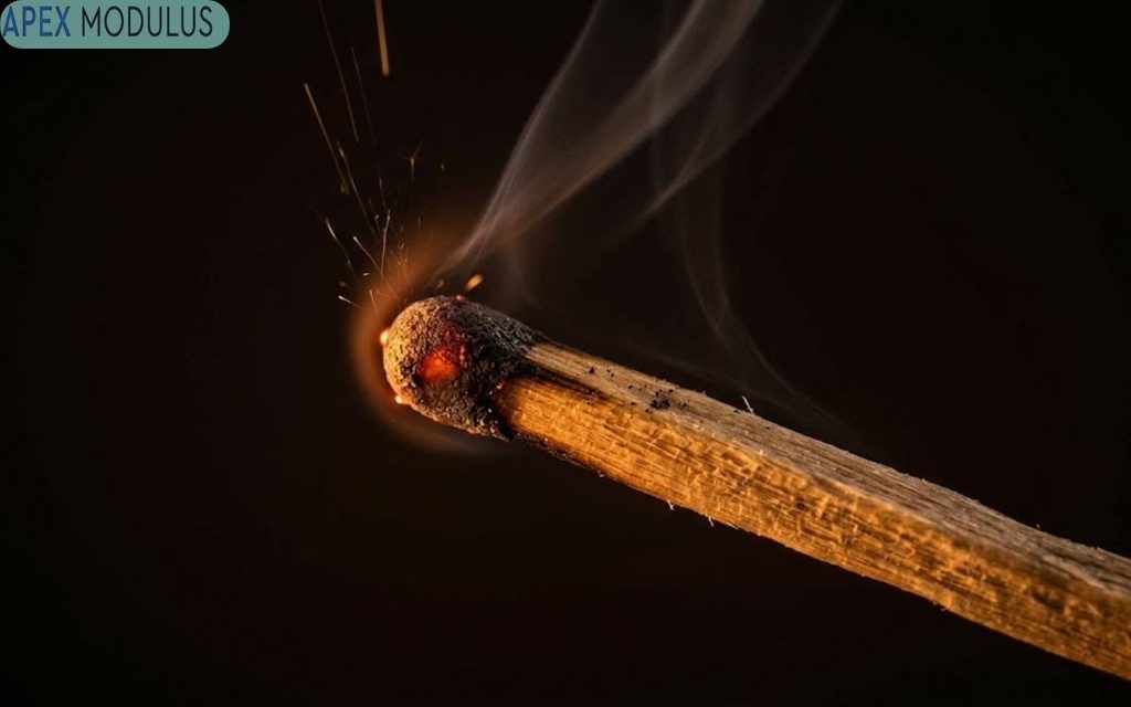 A close-up photograph of a just-lit matchstick head, showing the glowing ember, sparks, and delicate wisps of smoke against a dark background.